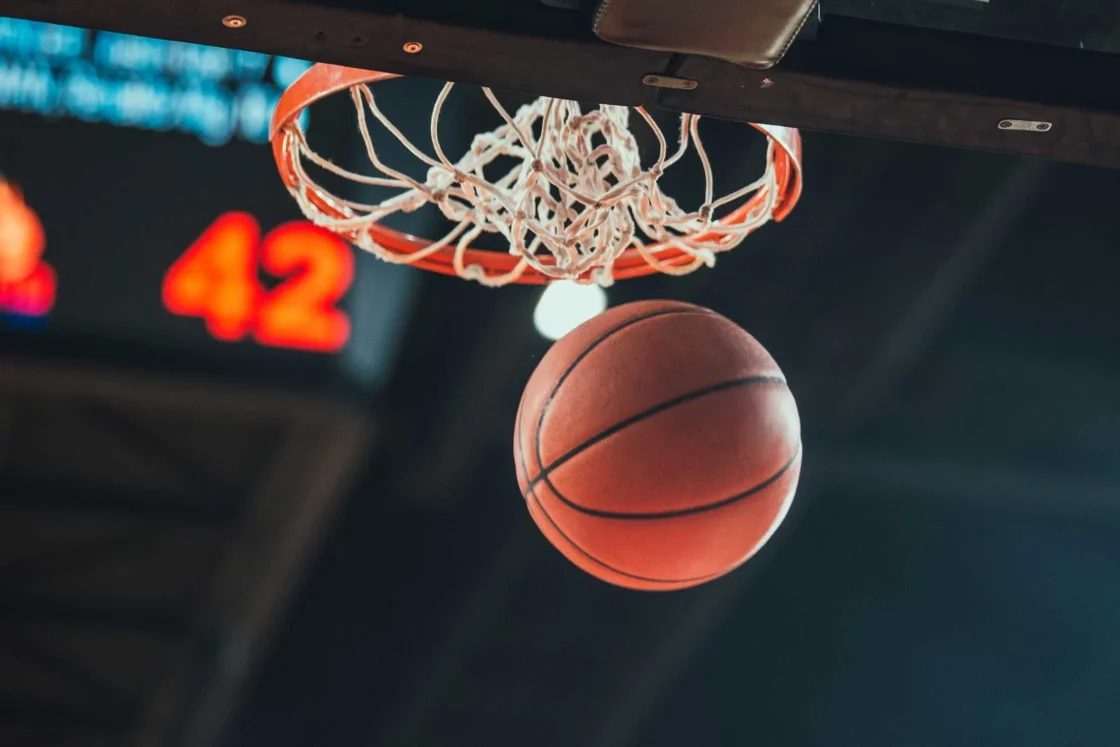 Basketball going through hoop with blurred scoreboard in background.