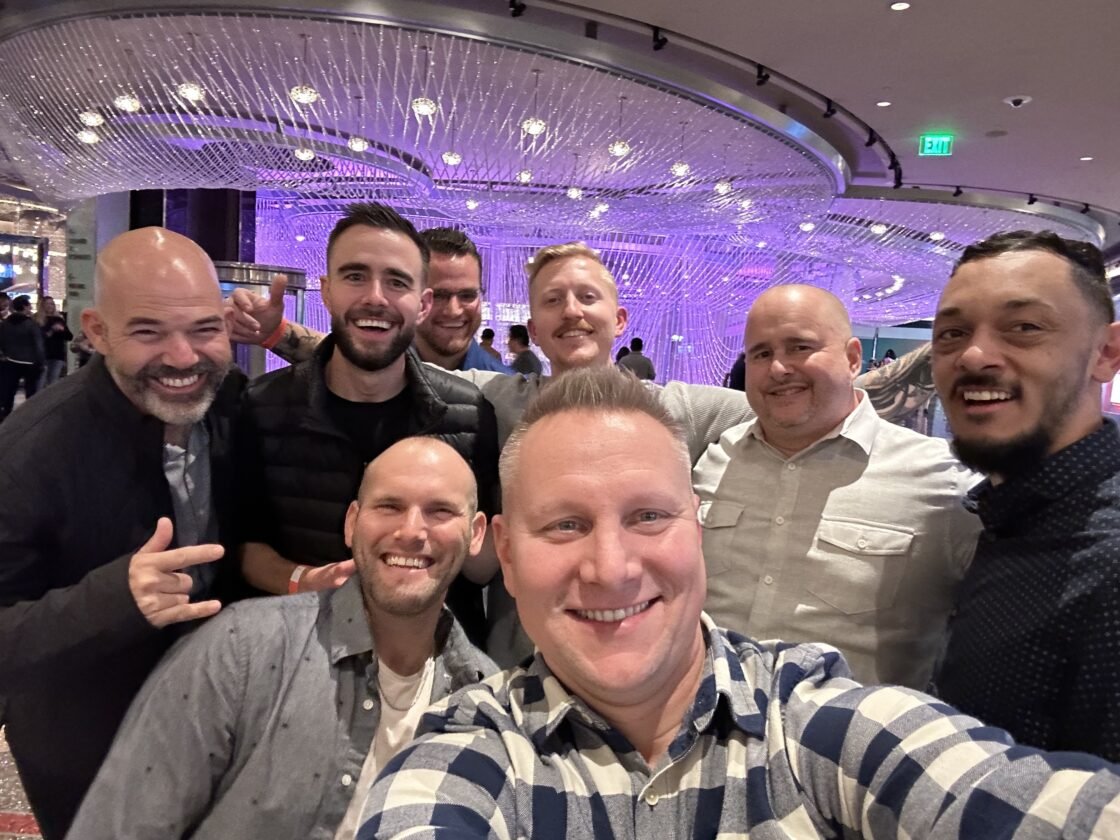 A group of eight men smiling for a photo in a modern, brightly lit room.