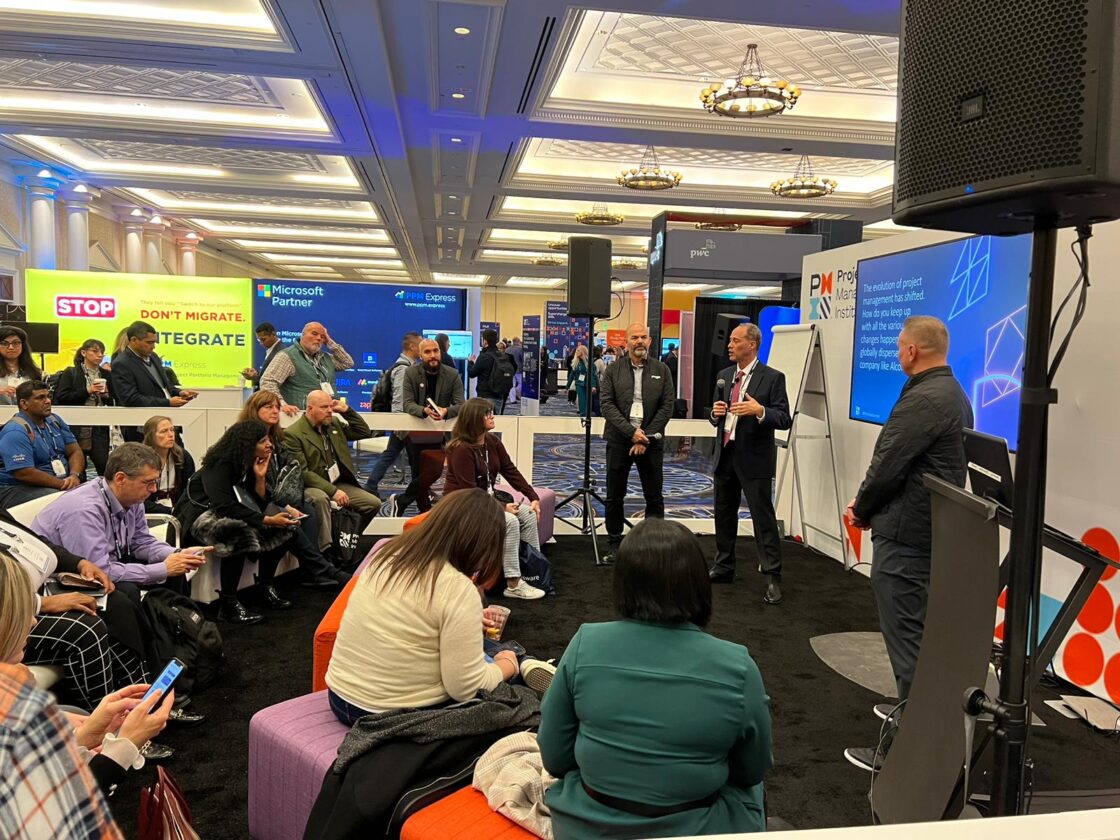 People attending a presentation at a tech conference with colorful booths in the background.