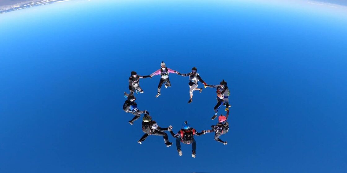 Skydivers forming a circle mid-air against a clear blue sky backdrop.