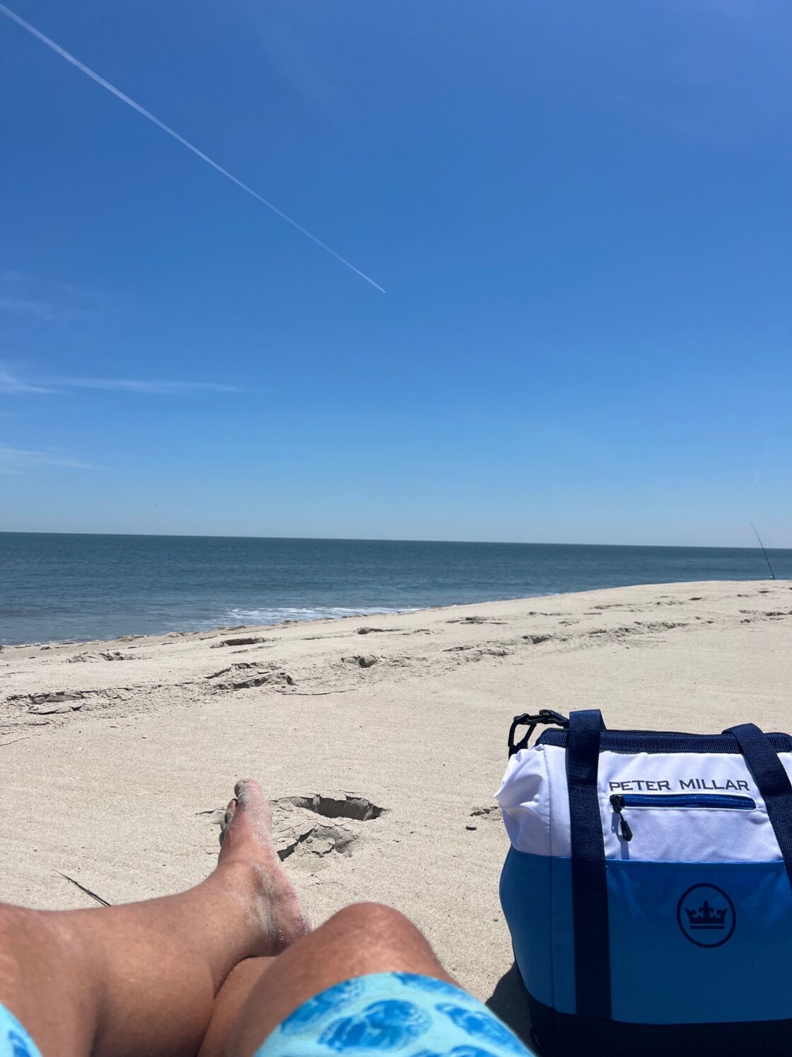 Person relaxing on a sandy beach with a blue cooler bag nearby, ocean in the background.