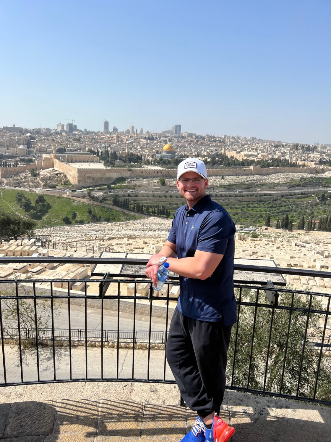 Man in casual wear smiles near a scenic overlook of a city skyline and historic site.