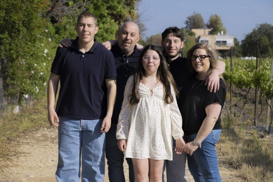 A family of five posing outdoors with trees and a house in the background.