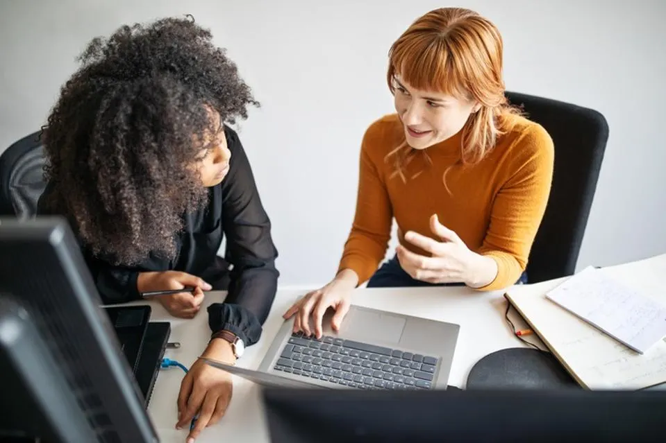 Two women discussing and working on a laptop at a desk with notebooks.