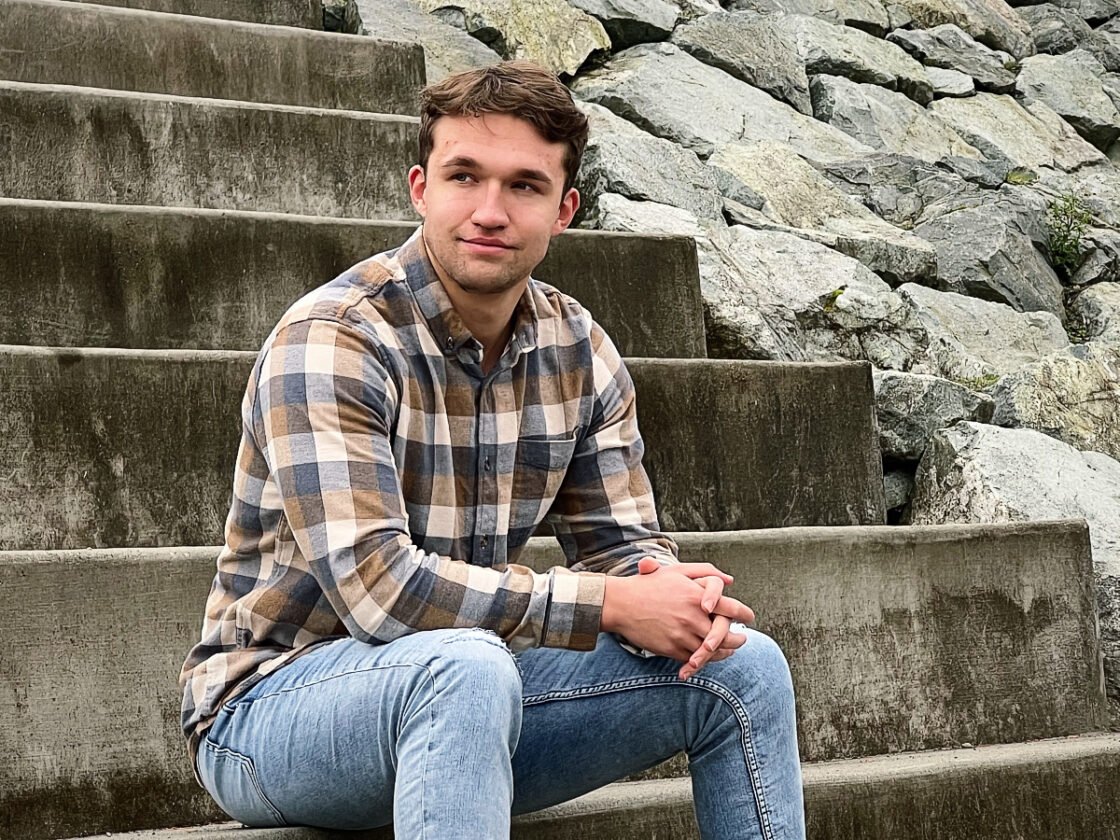 Man in plaid shirt sitting on stone steps near a rocky wall.