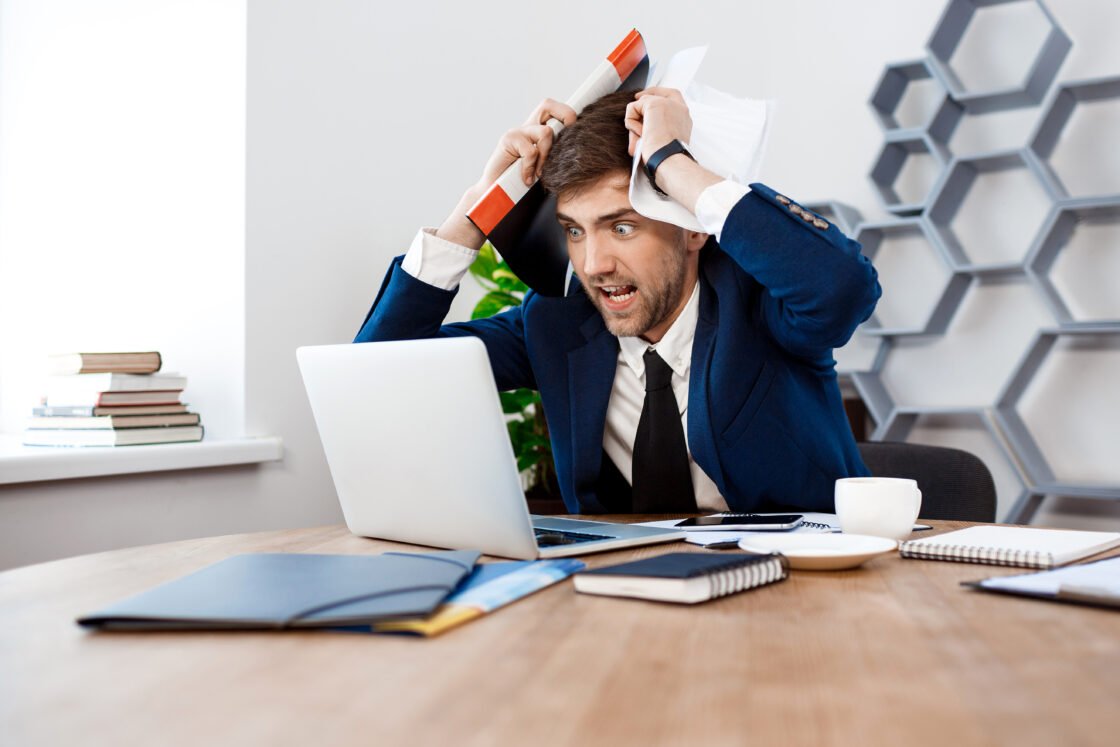 Man in suit looking stressed at laptop, holding papers above head, sitting at a desk.