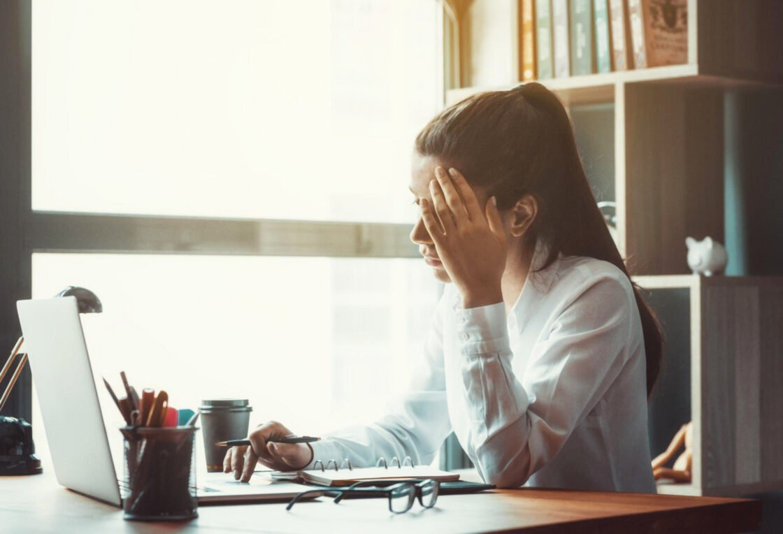 A woman in a white shirt looks stressed while working on a laptop in a sunlit room.