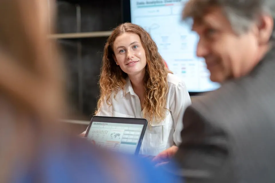 A woman in business attire smiles during a meeting, with a laptop in front of her.