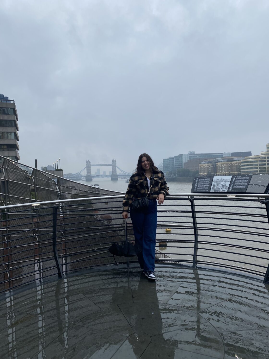 A person standing on a riverside walkway with Tower Bridge in the background on a cloudy day.