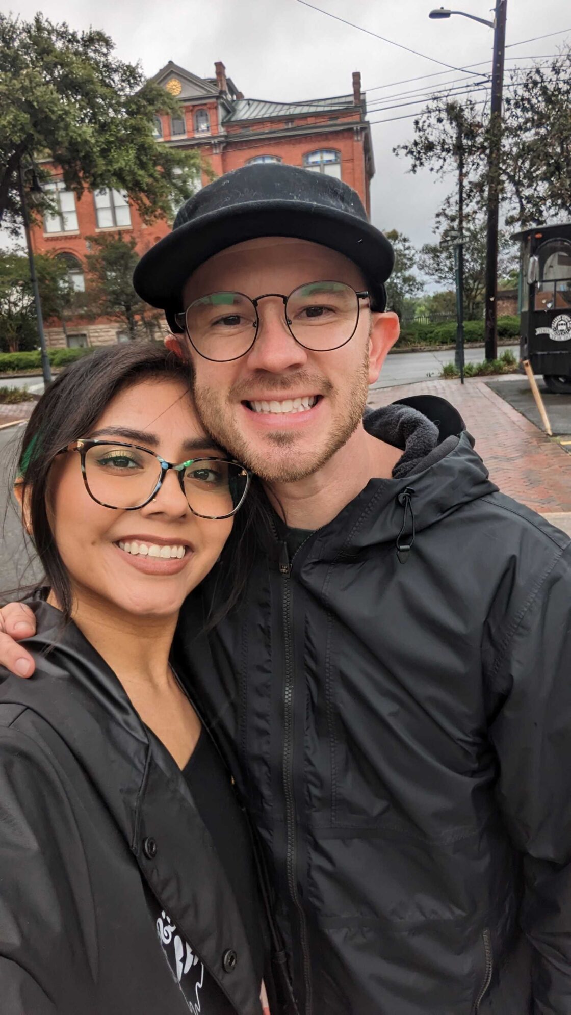 A smiling couple in glasses taking a selfie outdoors on a cloudy day.