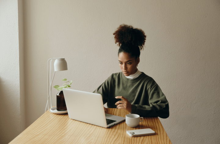 Person using a laptop at a wooden desk with a plant, small lamp, phone, and cup.