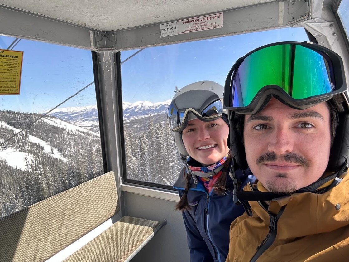 Two people in ski gear on a gondola with snowy mountains in the background.