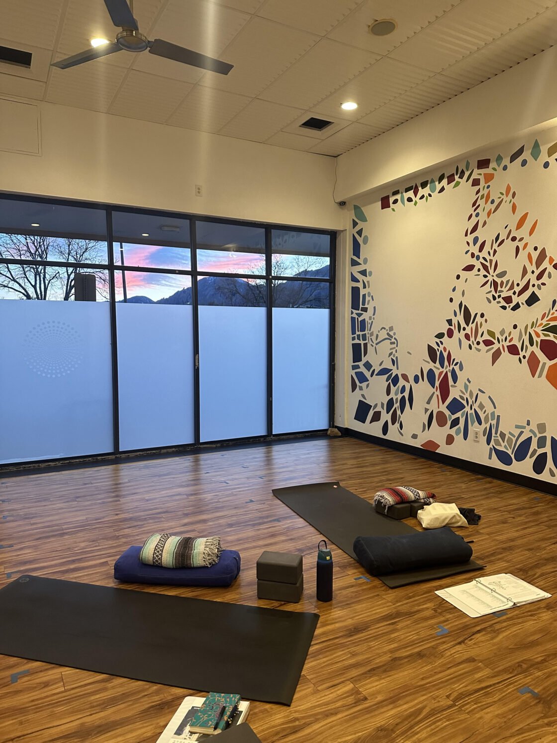 Yoga studio with mats, cushions, and a colorful wall mural; sunset visible through windows.