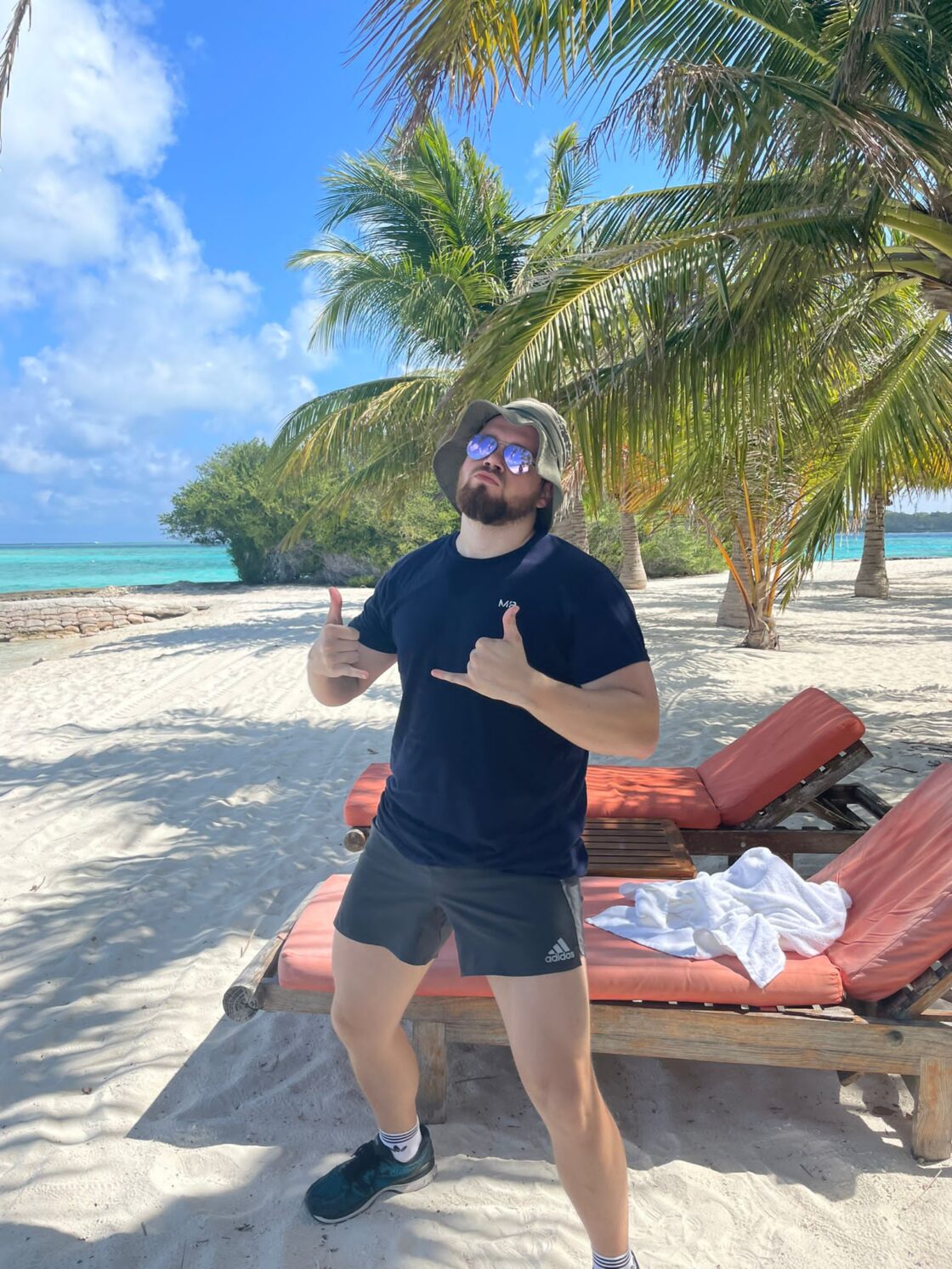 Man on a tropical beach in casual attire, posing under palm trees near red lounge chairs.