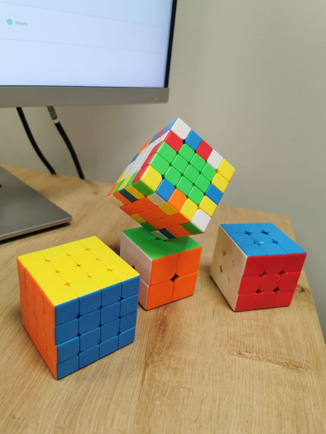 Four colorful Rubik's cubes stacked on a wooden desk near a monitor.