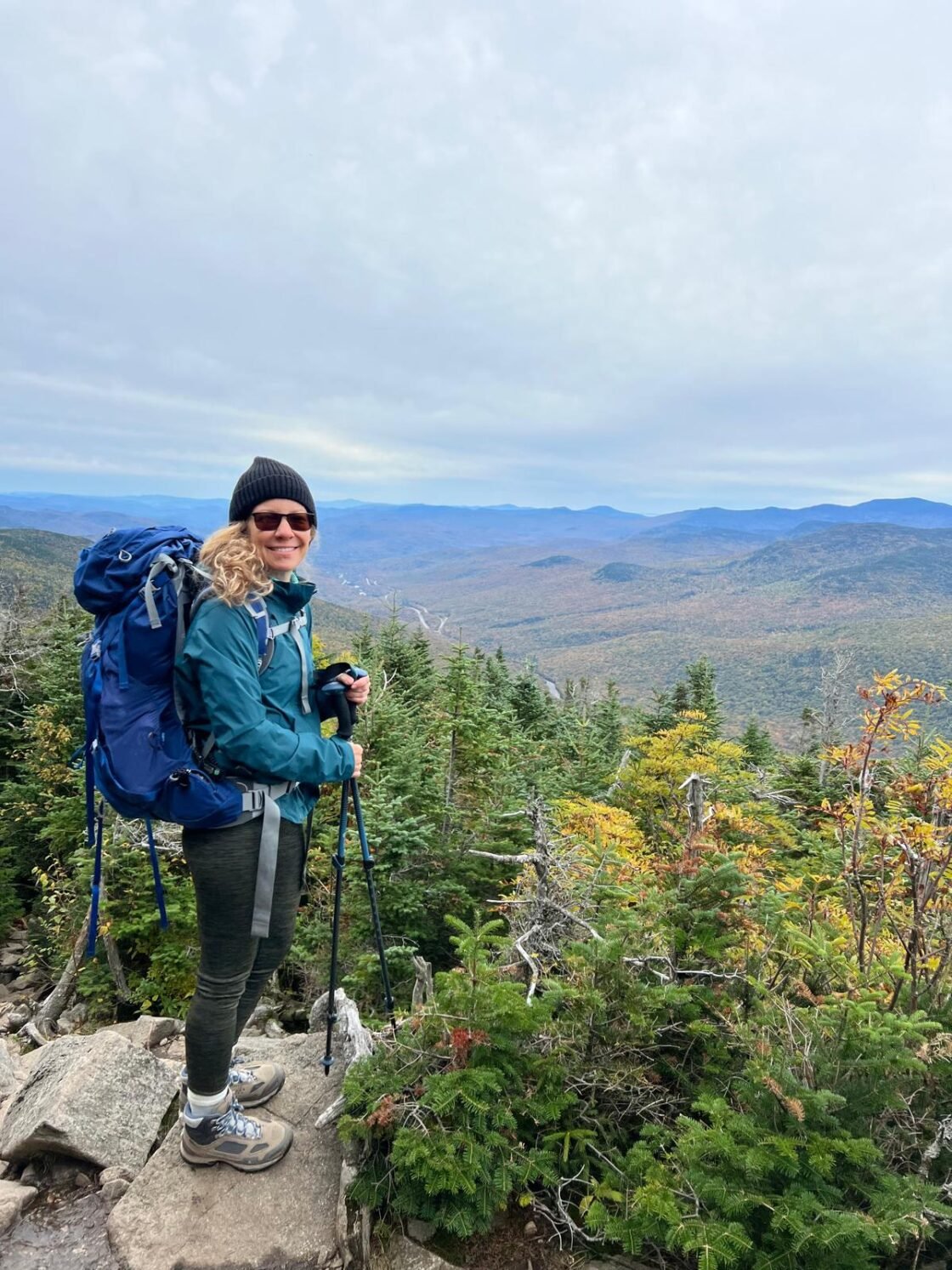 A woman hiking, standing on a rock, overlooking a vast, forested mountain landscape.